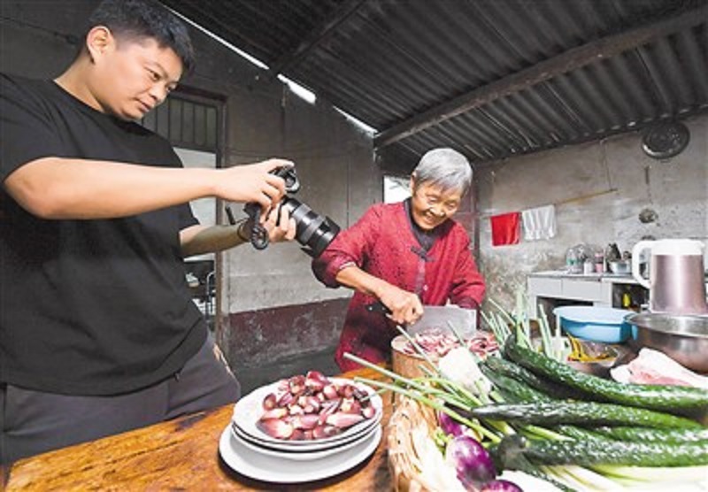 張萬露是一名返鄉(xiāng)創(chuàng)業(yè)的短視頻博主，用鏡頭記錄生活在陜西省安康市漢濱區(qū)棗園村的爺爺奶奶家里日常生活，吸引了200多萬名粉絲，并通過直播帶貨讓網(wǎng)友品嘗“家的味道”。圖為張萬露（左）在廚房拍攝奶奶切菜。鄒競一攝（新華社發(fā)）